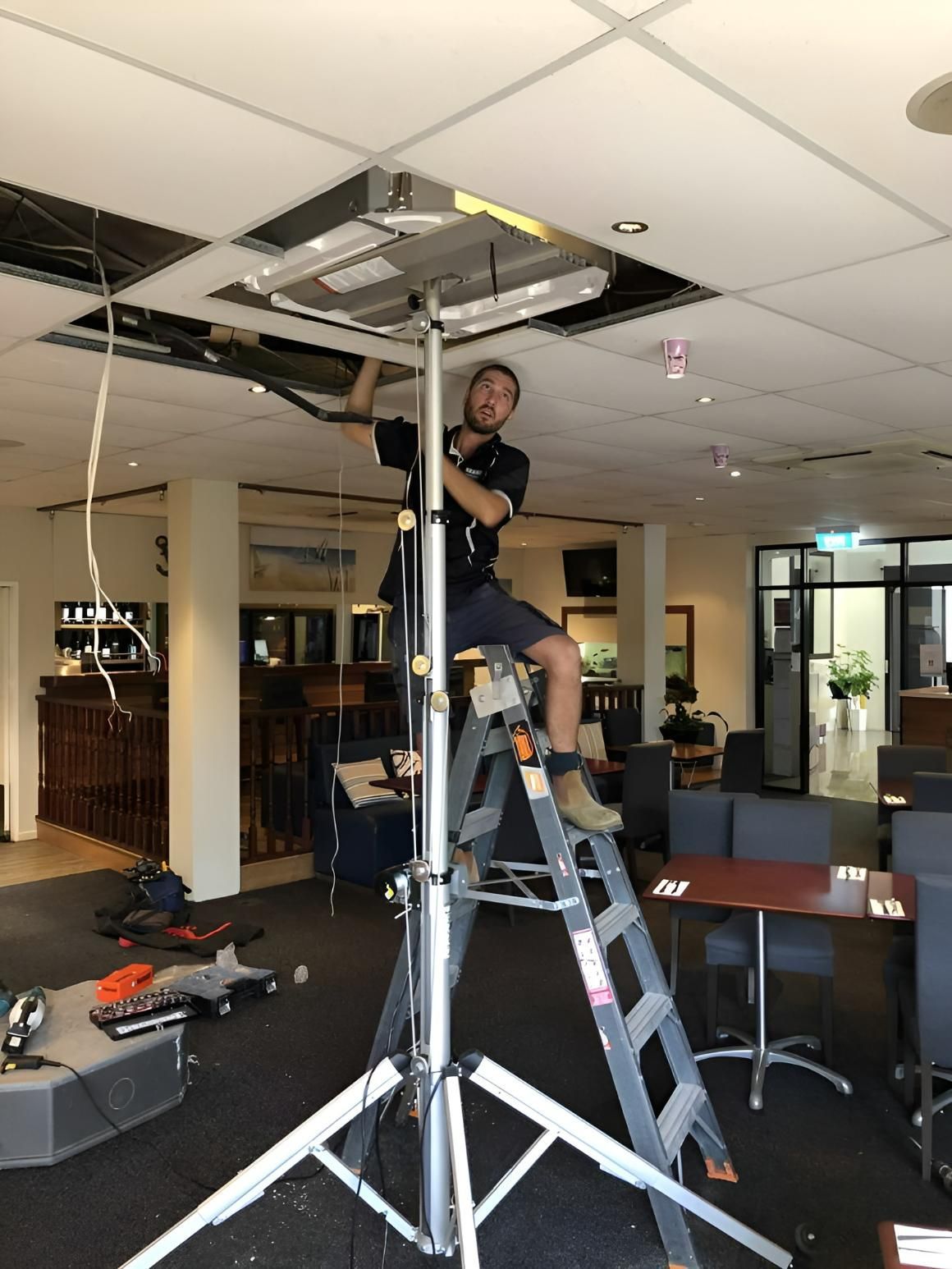 A Man Is Sitting On A Ladder Working On A Ceiling Fan — Beards Refrigeration & Air Conditioning in Bucasia, QLD