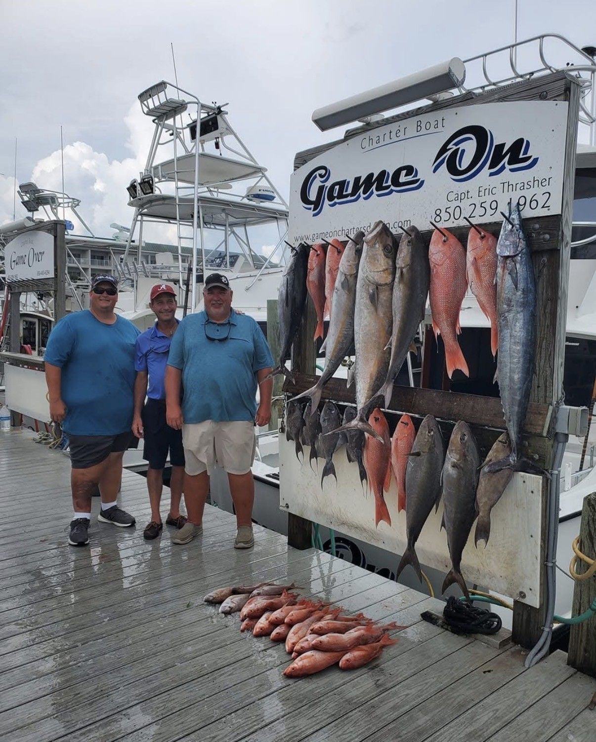 Two men standing by a dockside fish display with many hanging fish and fish laid on the ground.