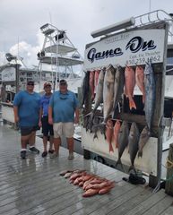 Three men stand by a dockside fish display with hanging fish and fish laid on the wet pier.