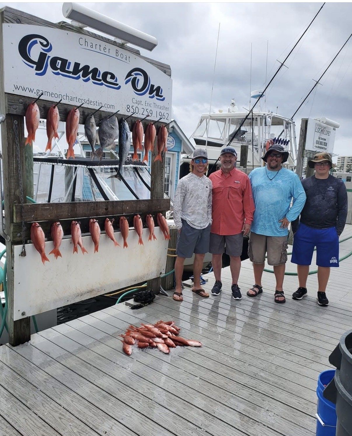 Four people pose beside a dockside fish display with hanging fish and a “Game On” sign.