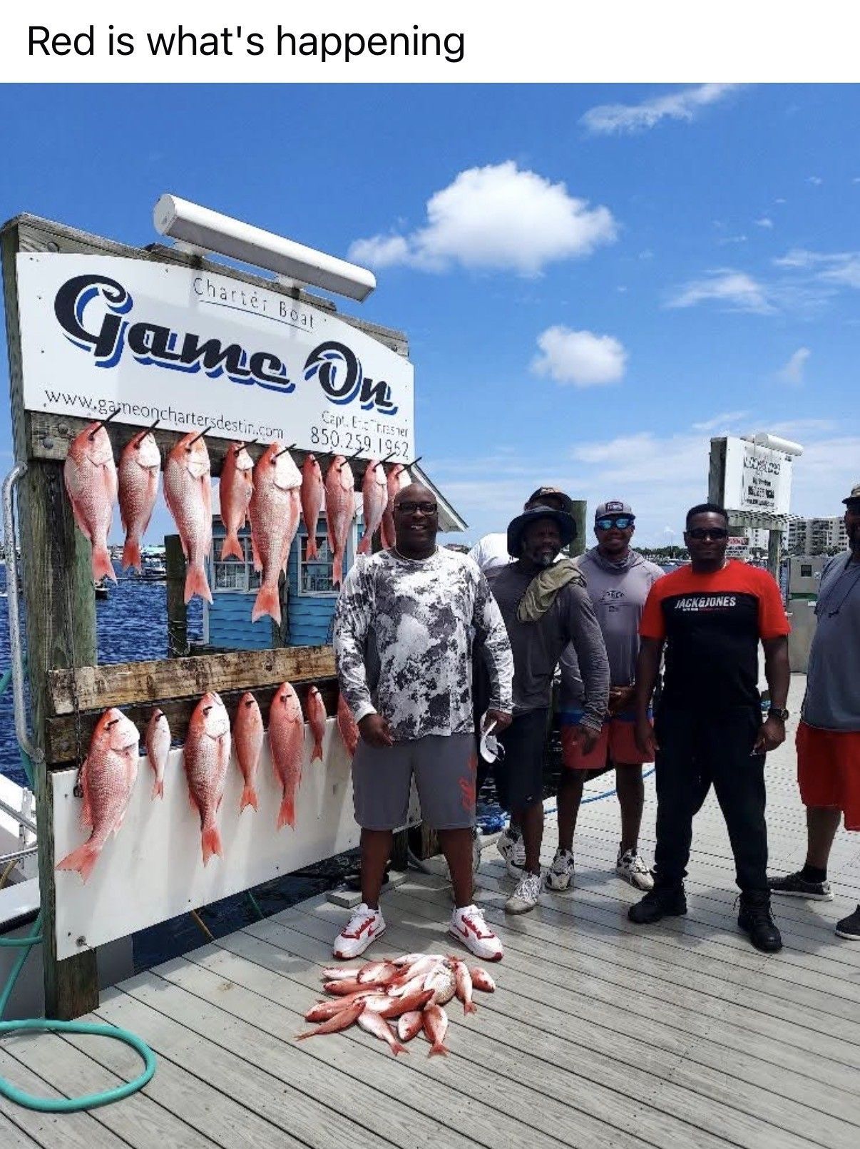 Three men pose beside a fish display and sign on a sunny boardwalk, with fish hanging and on the ground.