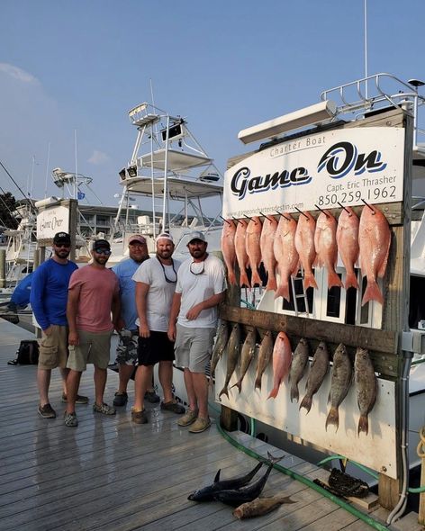 Five anglers posing beside a “Game On” sign with a long row of hanging fish on a dock at sunset