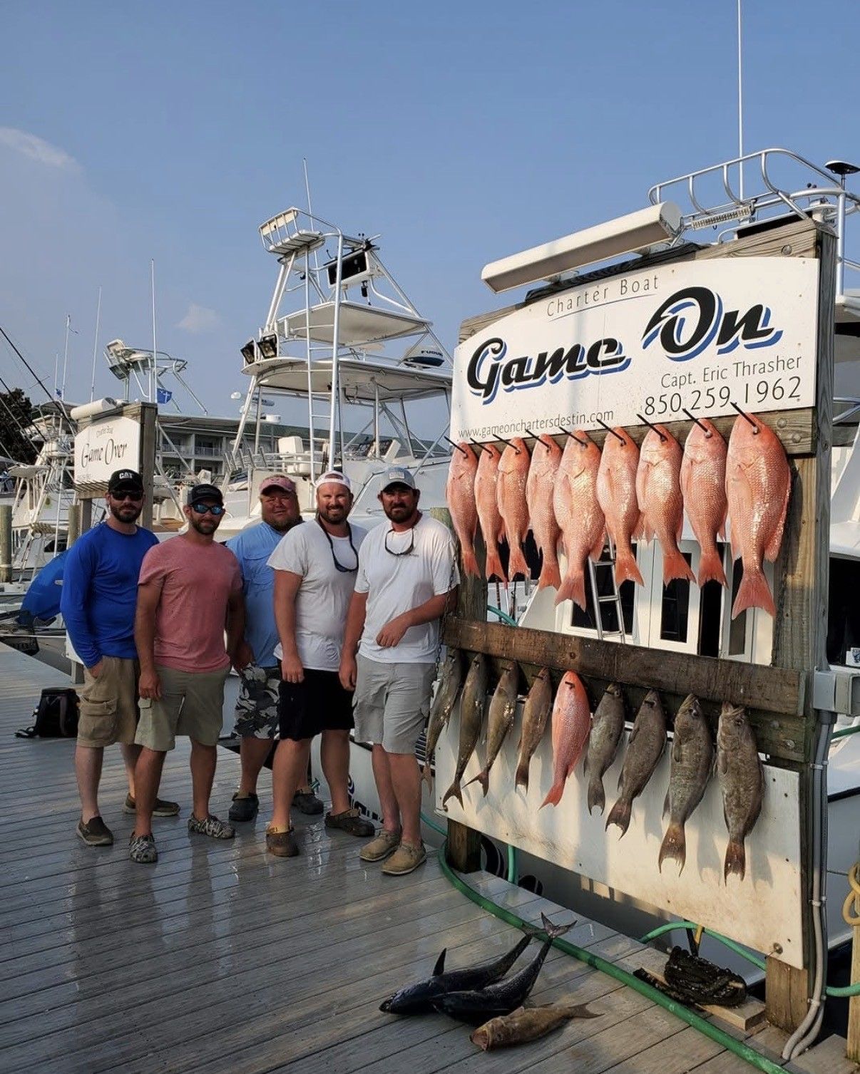 Five anglers pose by a fish rack on a dock, with a boat and marina in the background.