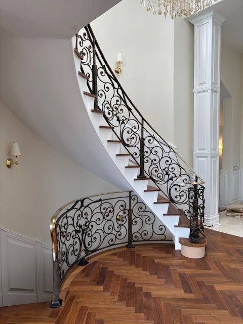 Curved staircase with ornate black wrought iron railing, white steps, and herringbone wood floor.