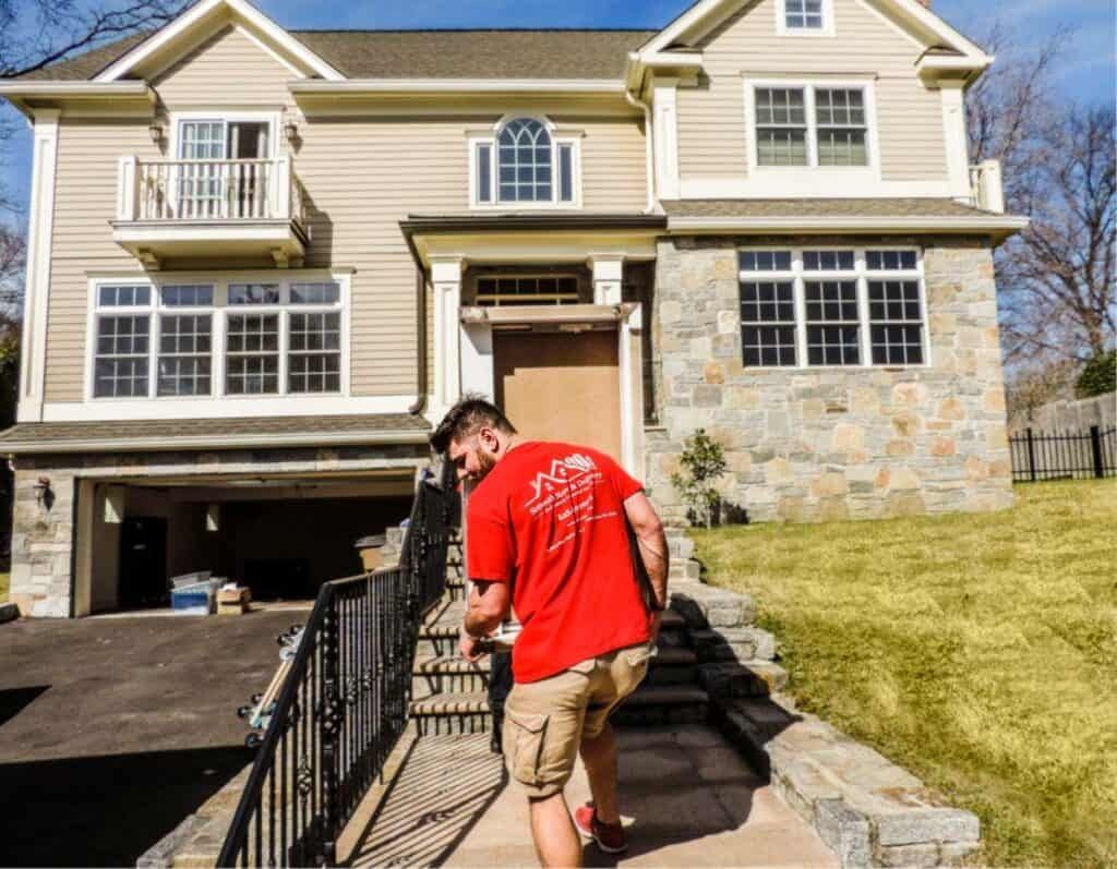 in the background, we see a beautiful house with a garage door open and a grass yeard on the right. in the center of the frame, smooth move and delivery movers wearing red branded shirts are carring a big heavy box