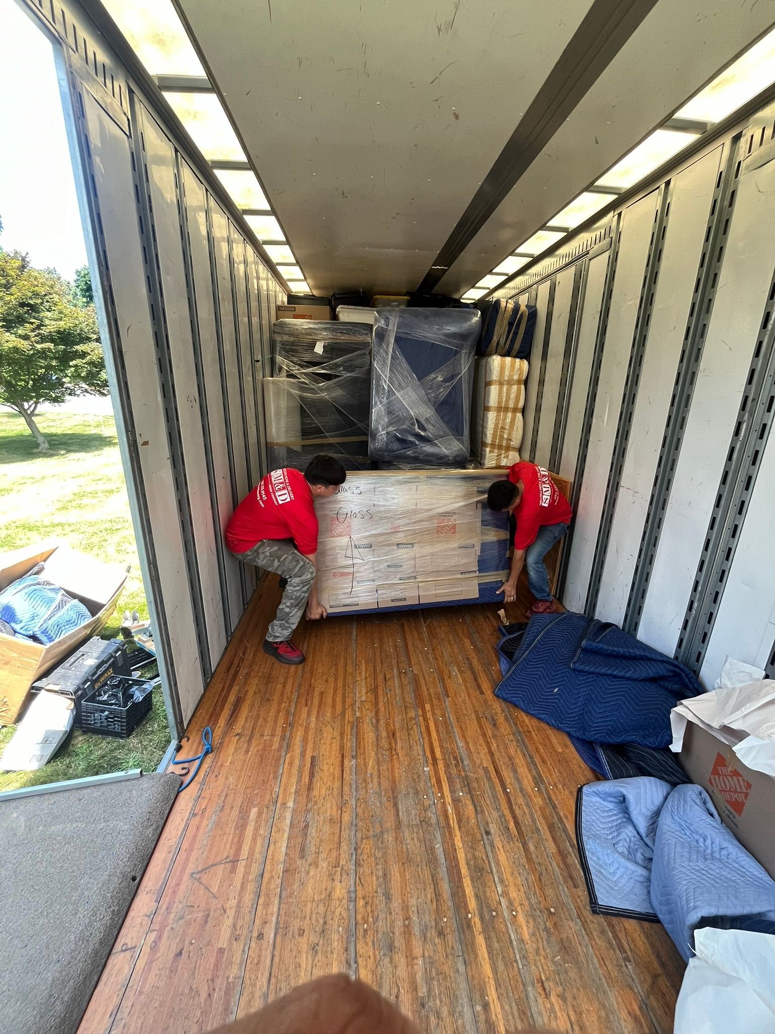 Two movers loading a trailer with packed furniture, boxes, and wrapped items.