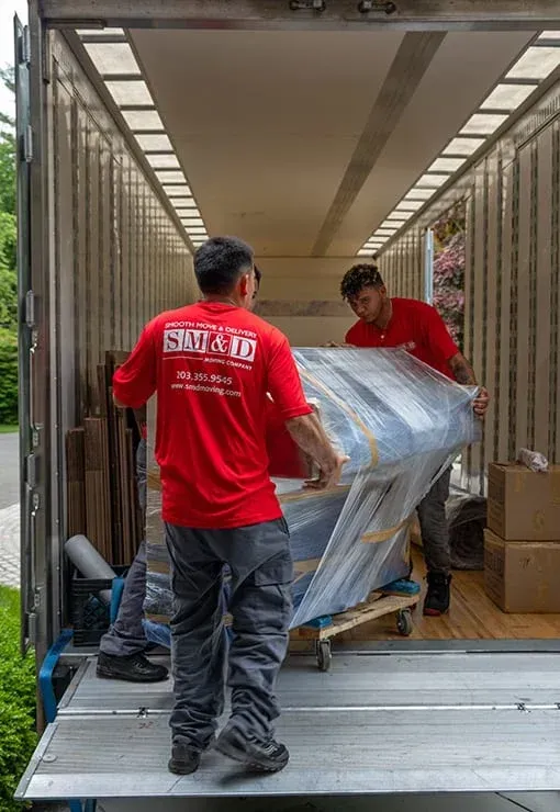 Two movers in red shirts loading furniture wrapped in plastic into a moving truck.