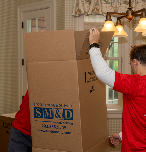 Movers in red shirts packing a large cardboard box inside a brightly lit room.