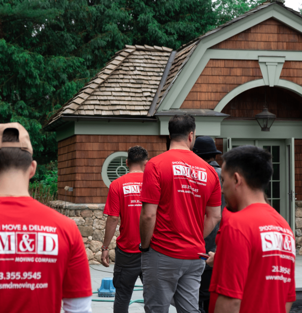 Movers wearing red shirts, standing outside a house with a brown shingle roof.