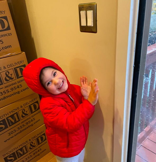 A young child in a red jacket smiles and claps near a light switch. Boxes are in the background.