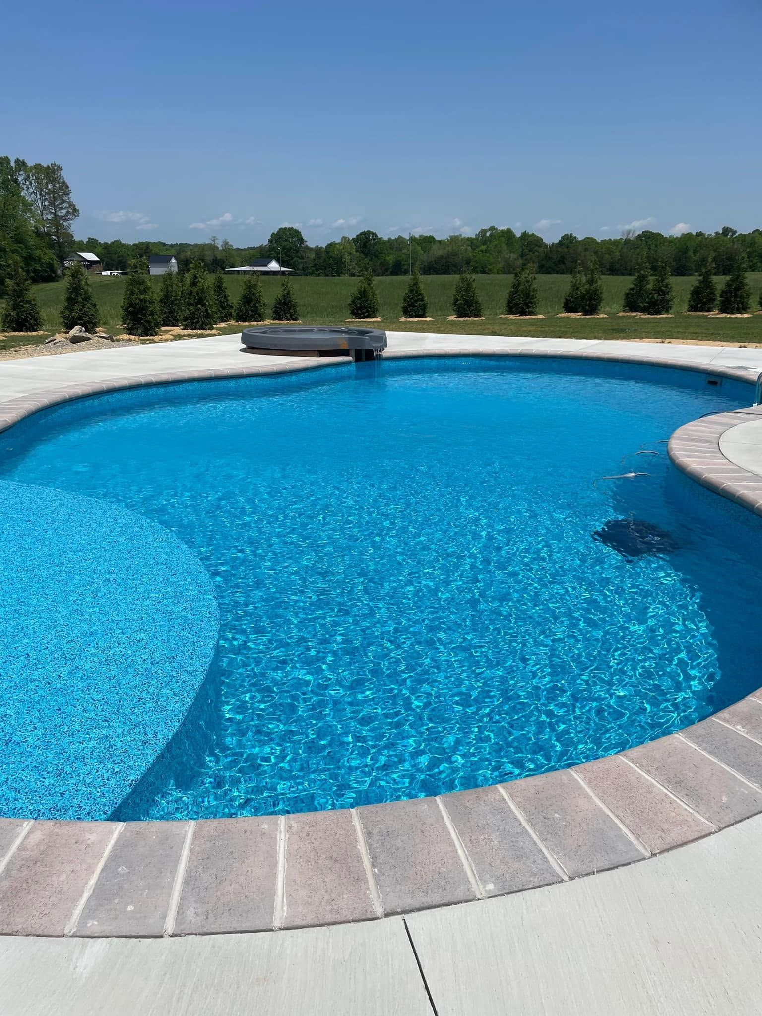 Curved blue swimming pool with stone coping and landscaped greenery under a clear sky