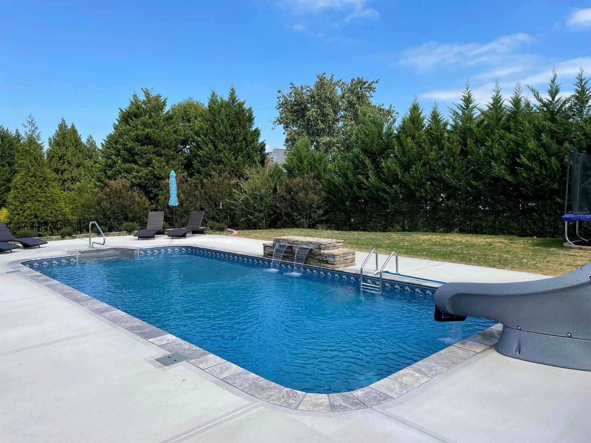 Rectangular outdoor swimming pool with blue water, stone edging, and trees under a clear sky