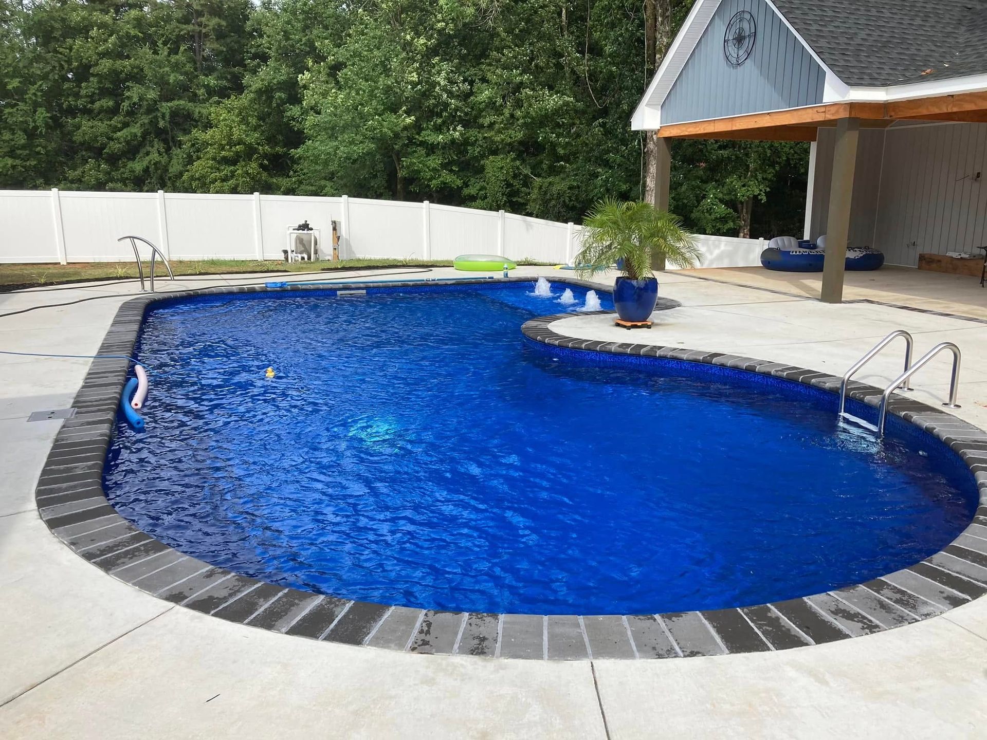 Curved blue backyard pool with white deck, stone trim, and potted plant beside a covered patio