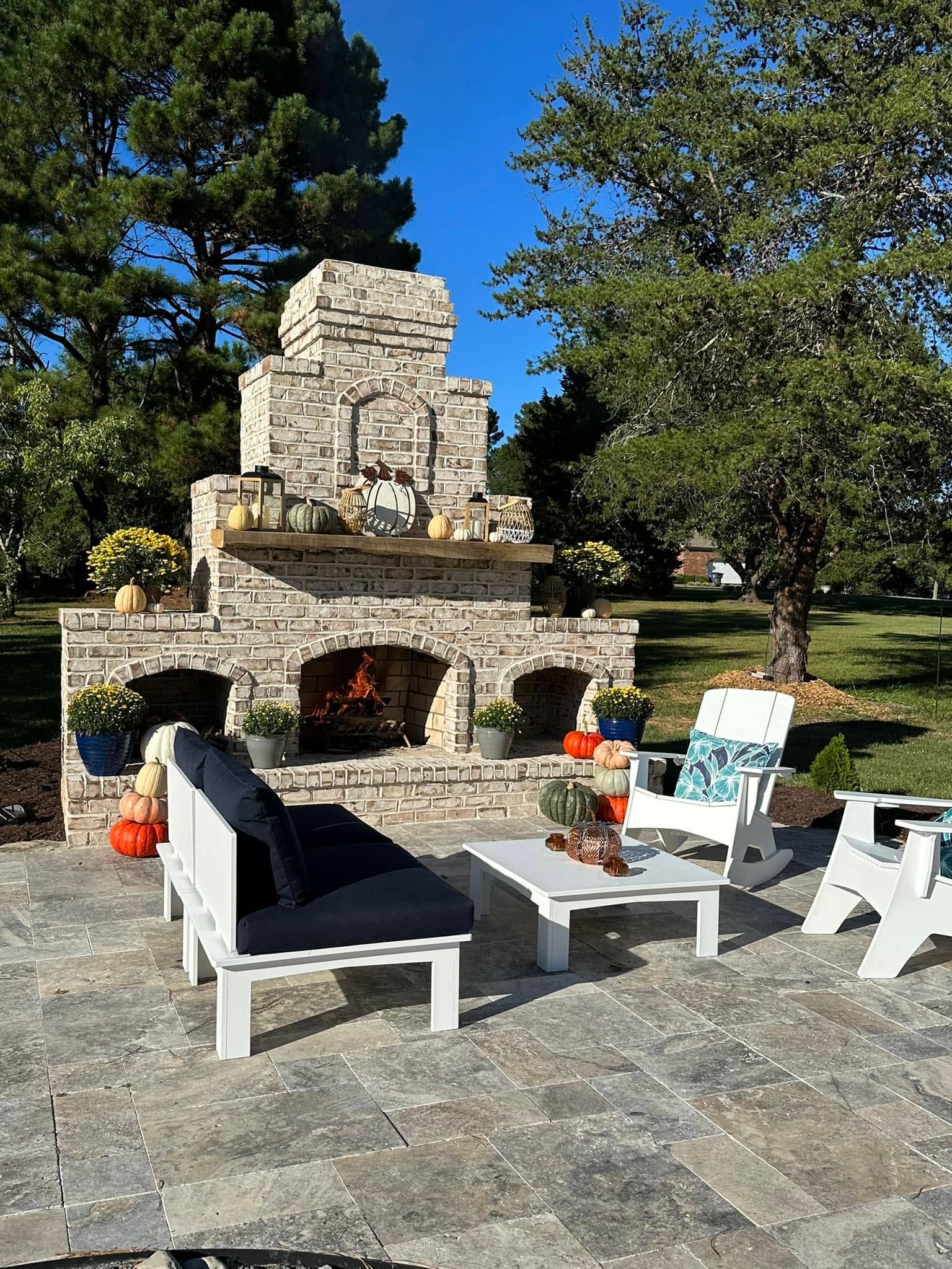 Stone outdoor fireplace with Adirondack chairs and benches on a patio, surrounded by trees and greenery
