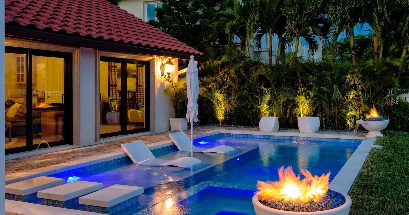 Luxury backyard pool at dusk with blue lighting, lounge chairs, and a fire pit beside a patio home