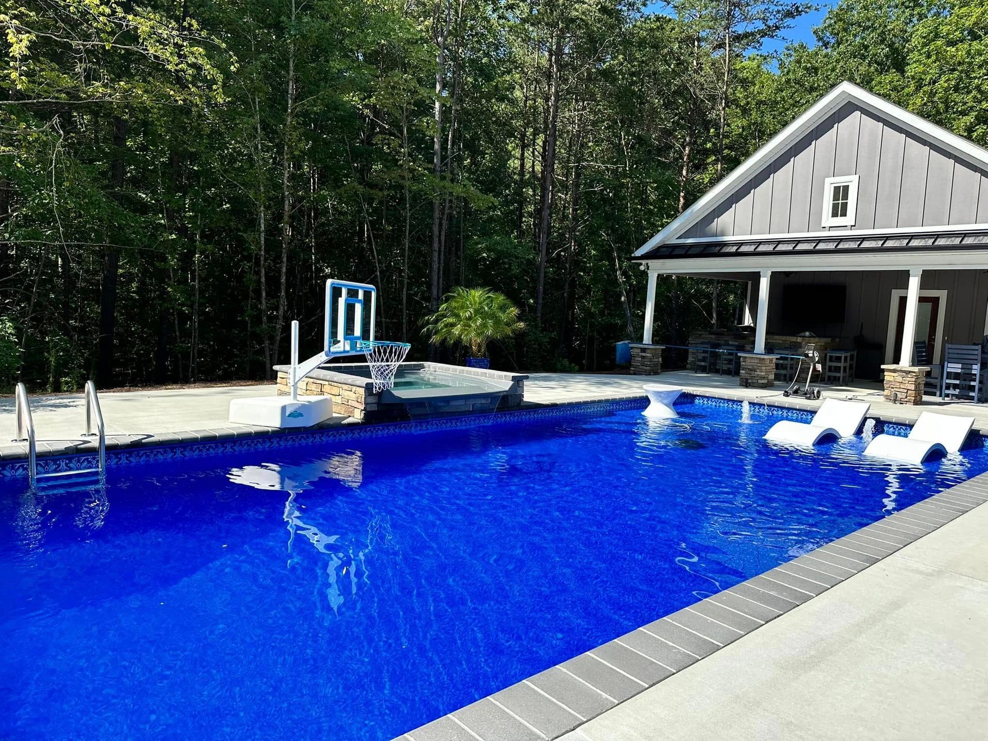 Backyard pool with bright blue water, white lounge chairs, and a gray house beside trees