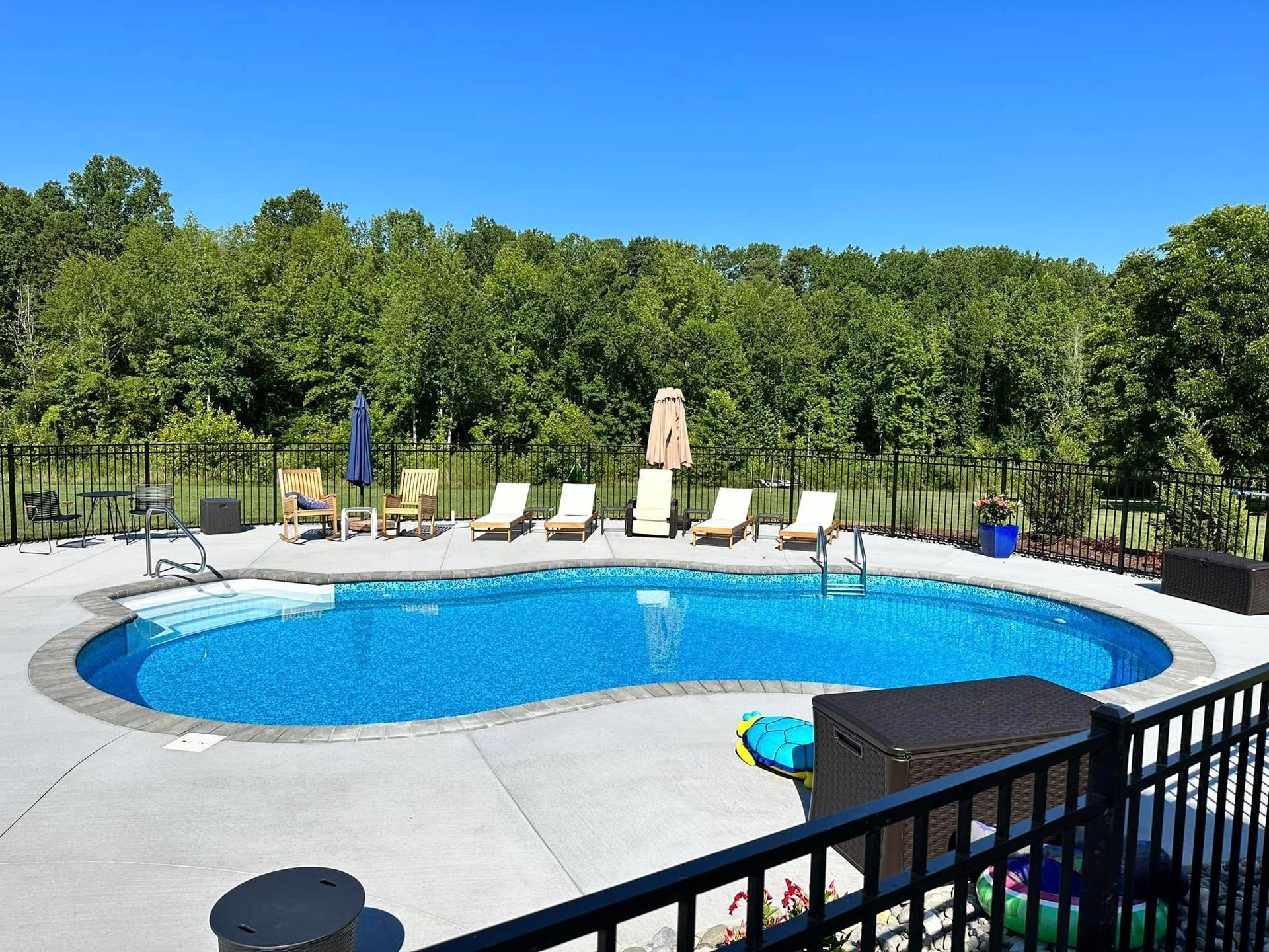 Backyard swimming pool with lounge chairs, blue umbrellas, and black fence overlooking trees