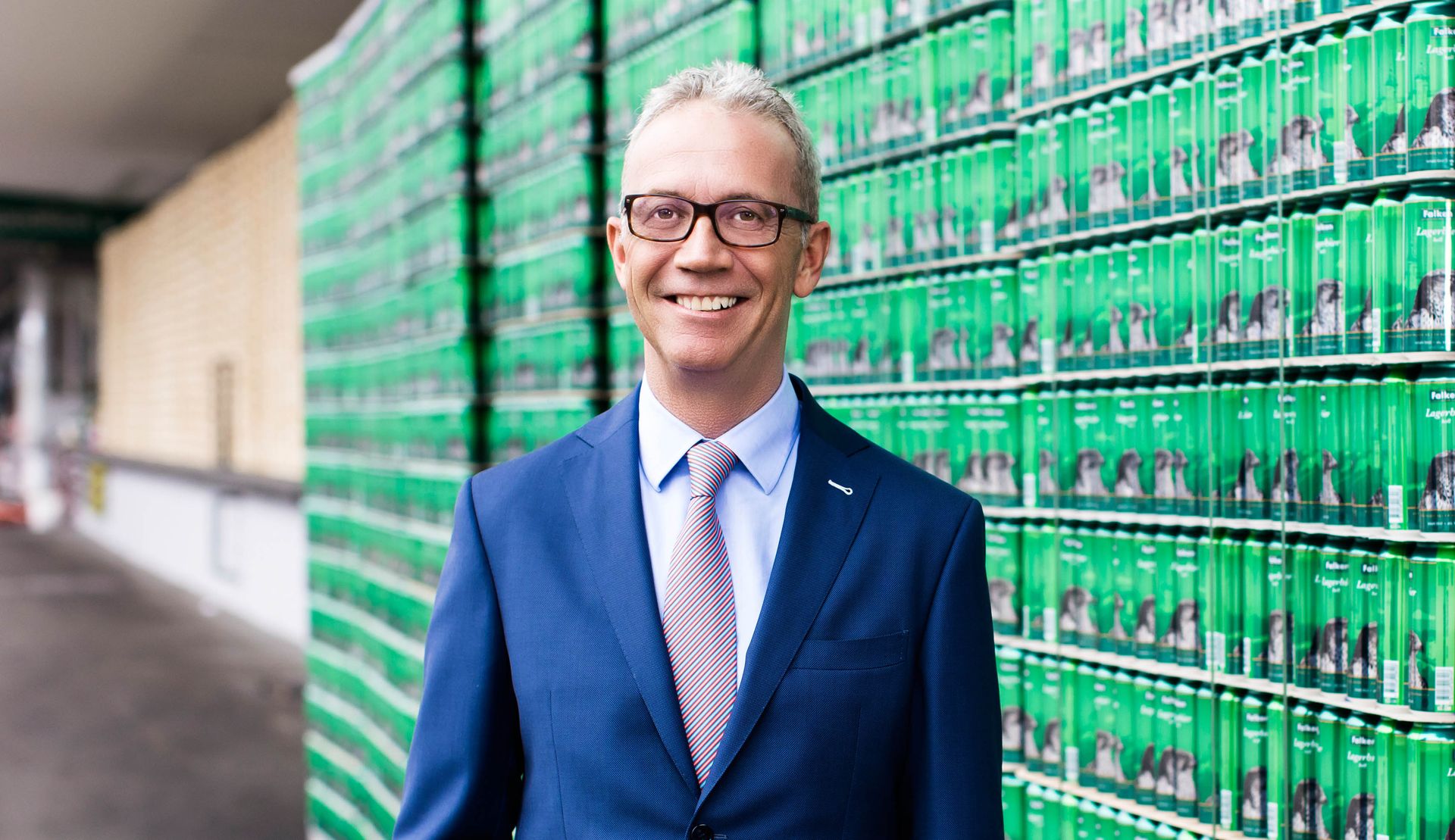 A man in a suit and tie is standing in front of a wall of green cans.