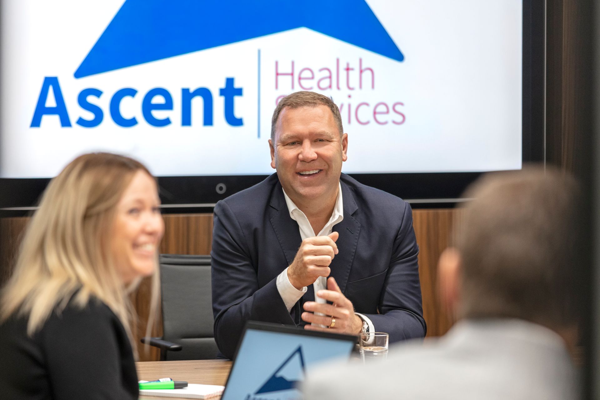 A group of people are sitting at a table in front of a screen that says ascent health services