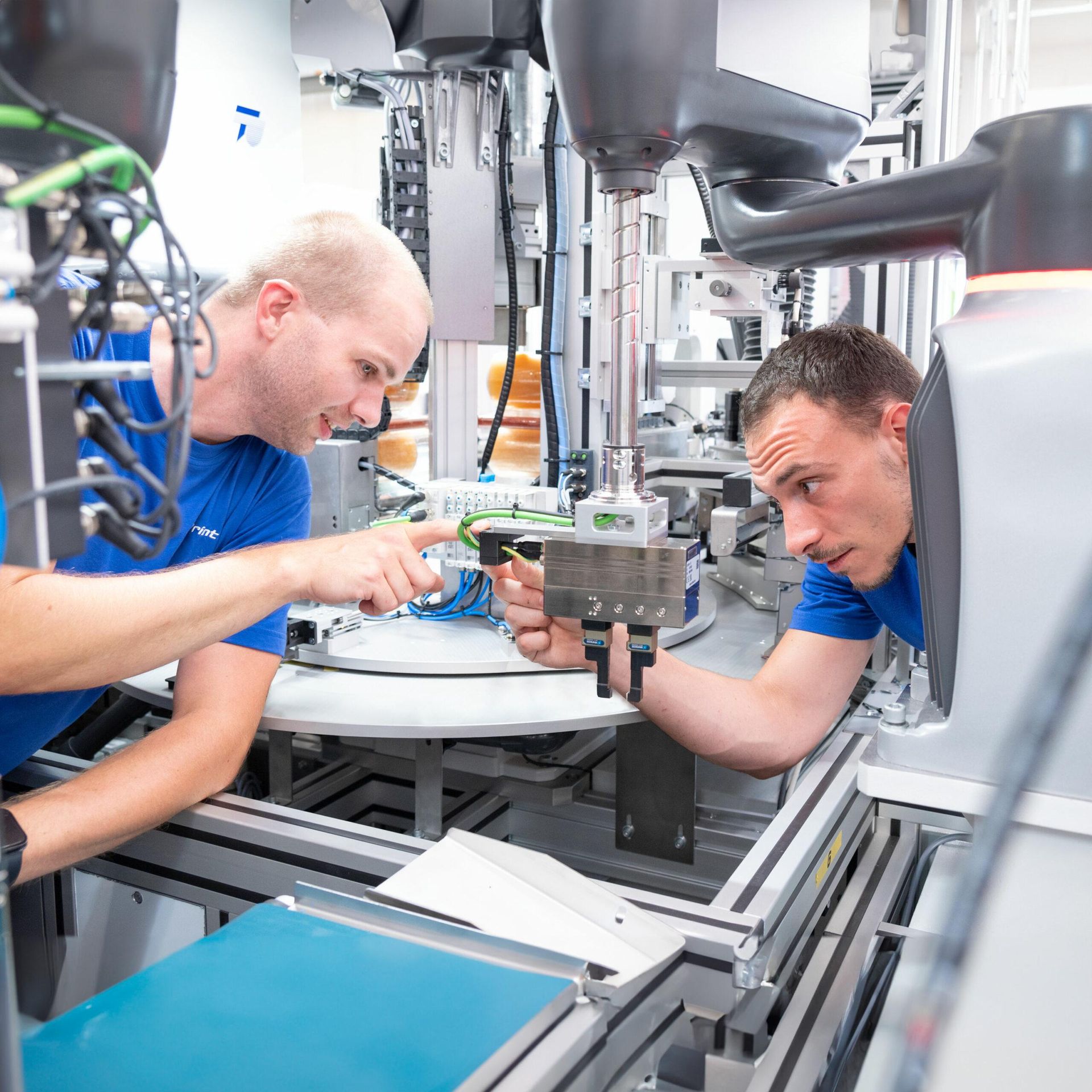 Two men in blue shirts examine machinery in a factory setting, one pointing.
