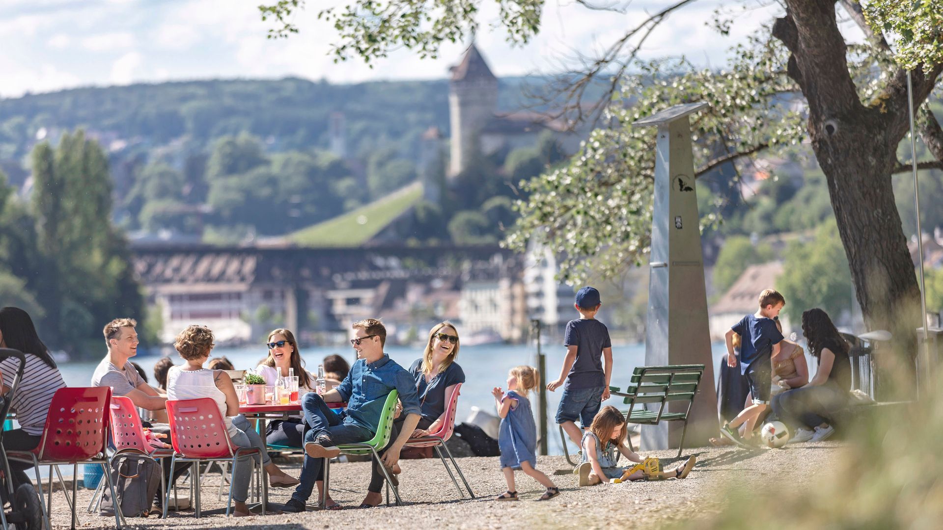 Eine Gruppe von Leuten sitzt an Tischen nahe des Rheins.