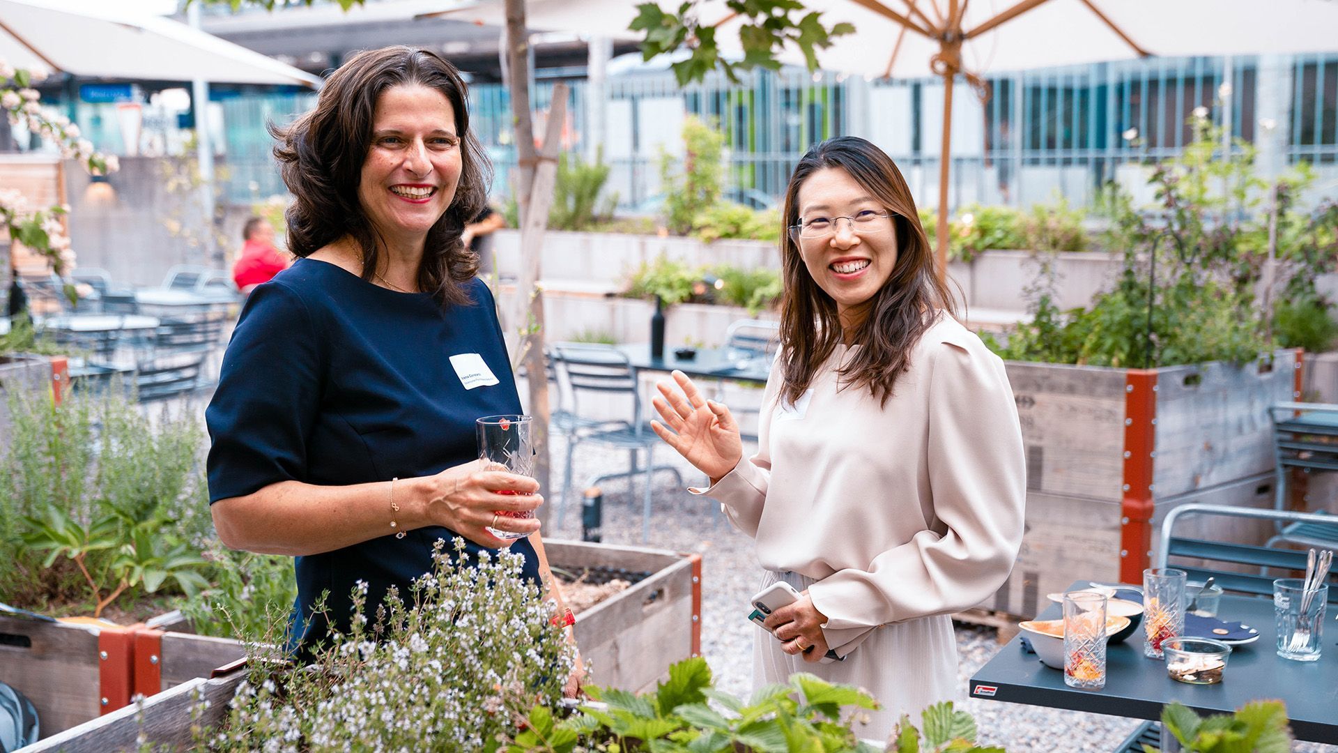 Two women are standing next to each other in a garden holding drinks.