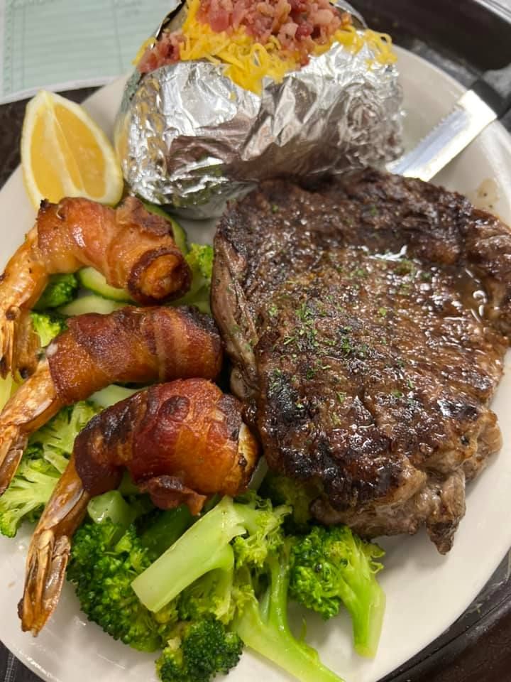 Grilled steak, shrimp, broccoli, and a foil-wrapped baked potato on a white plate with lemon wedge.