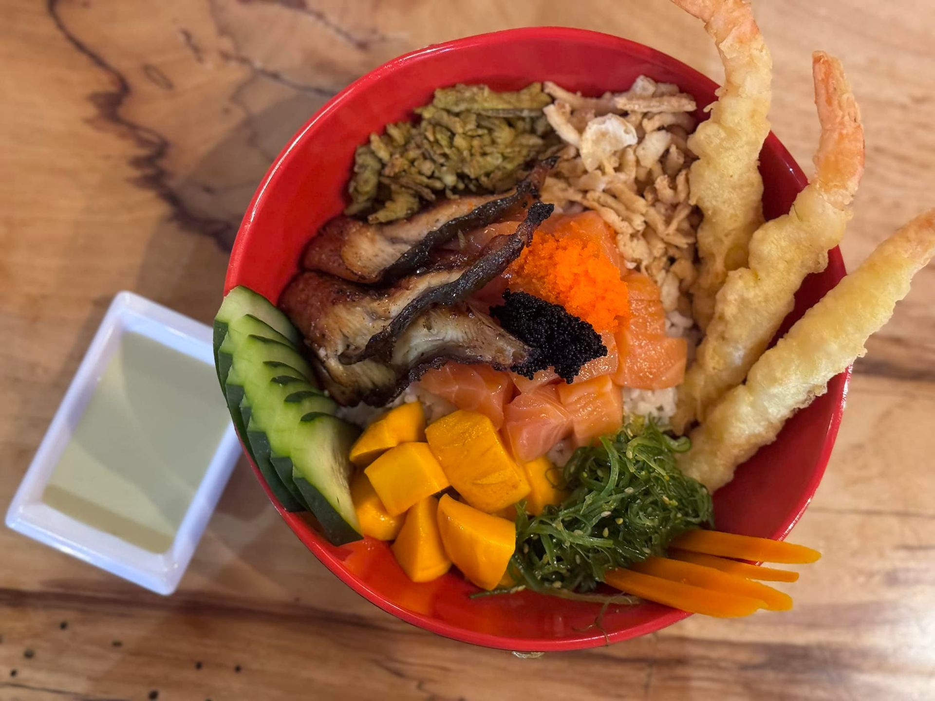 Sushi poke bowl with avocado, mango, salmon, vegetables, and tempura shrimp, plus a small sauce cup.