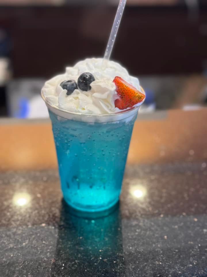 Blue drink topped with whipped cream, blueberries, and a strawberry in a clear plastic cup with a straw