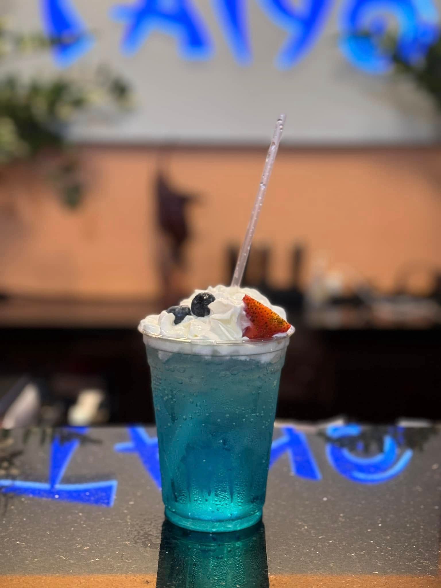 Blue cocktail with whipped cream and straw on a reflective bar counter, with blurred neon lights in the background