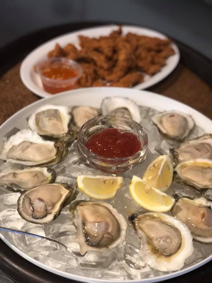 Oysters on ice with lemon wedges and cocktail sauce, with fried food in the background