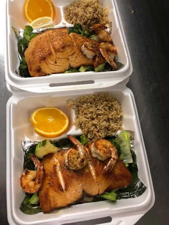 Two takeout trays with grilled chicken, rice, vegetables, and orange slices on the side.
