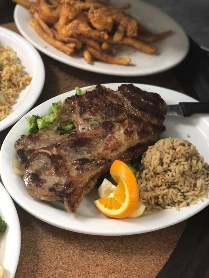 Grilled steak on a white plate with rice and an orange slice, with fried plantains and rice in the background