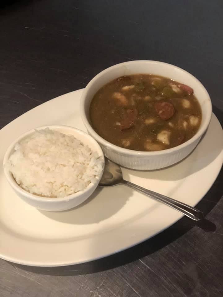 Rice and a bowl of stew with sausage on a white plate, with a spoon on a dark table.
