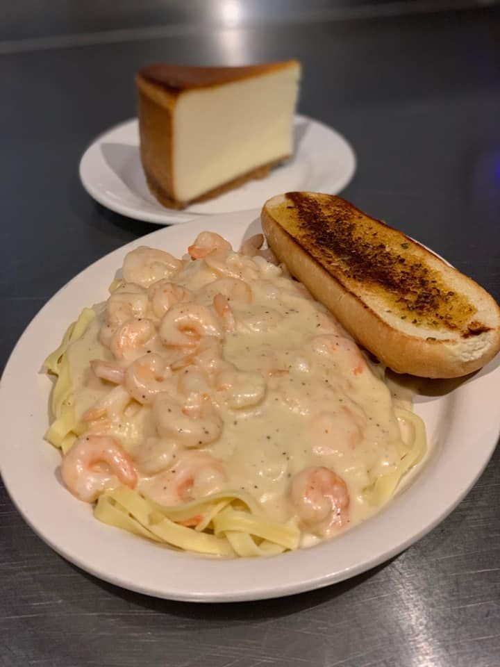 Plate of shrimp pasta with creamy sauce and garlic bread, with a slice of cheesecake in the background