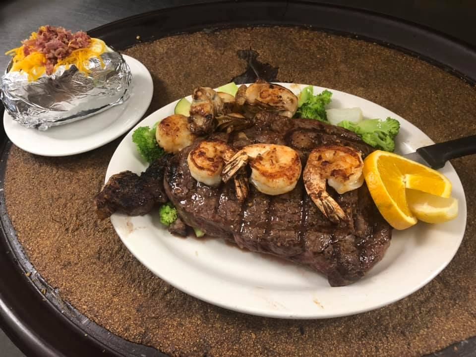 Grilled steak topped with shrimp and lemon wedges on a white plate, with a baked potato on the side.