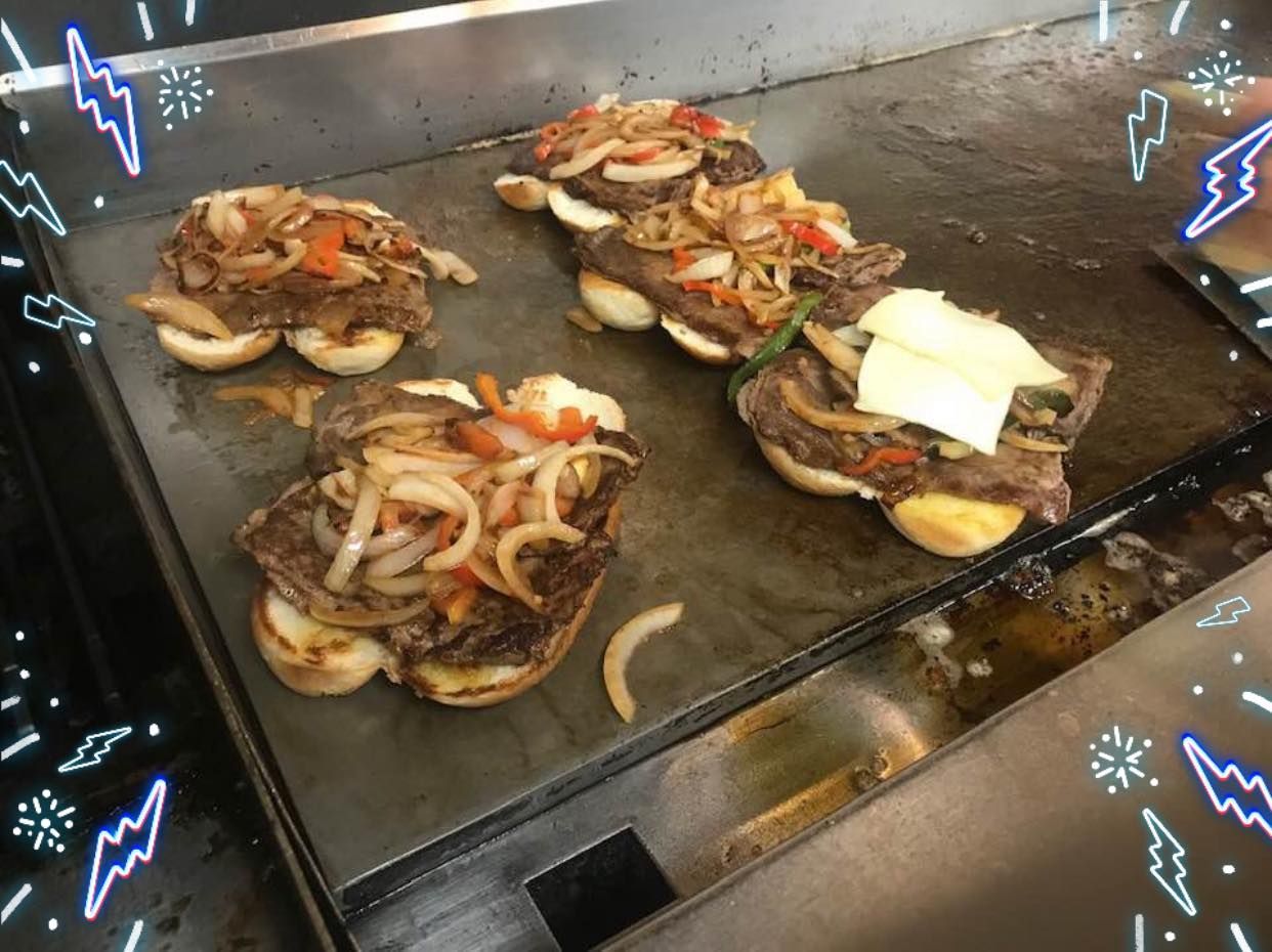 Hamburger patties cooking on a griddle, topped with onions, peppers, and cheese slices