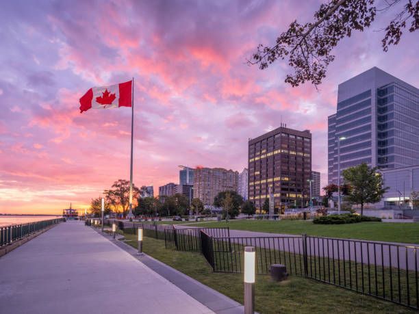 Canadian flag at sunset beside a waterfront path and modern city buildings under a pink sky