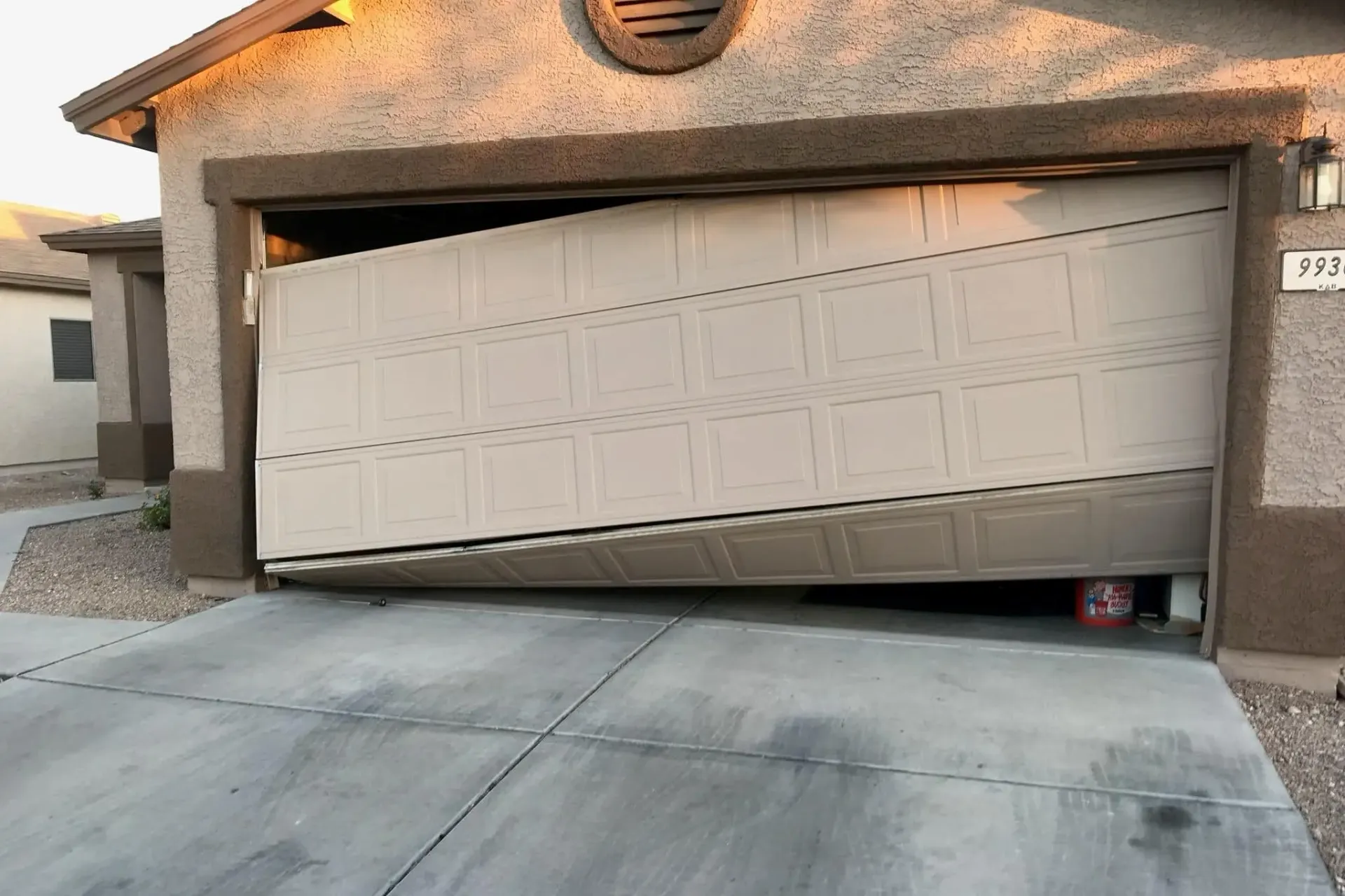 Damaged garage door, partially open and leaning. Beige door, tan trim, concrete driveway.