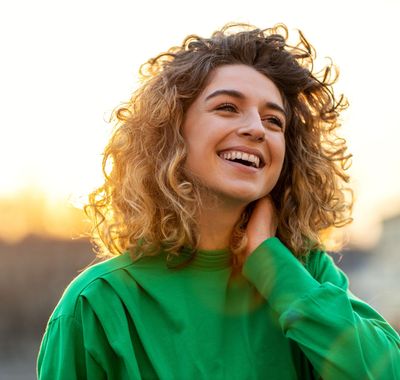 Smiling person in a green sweater with curly hair outdoors at sunset