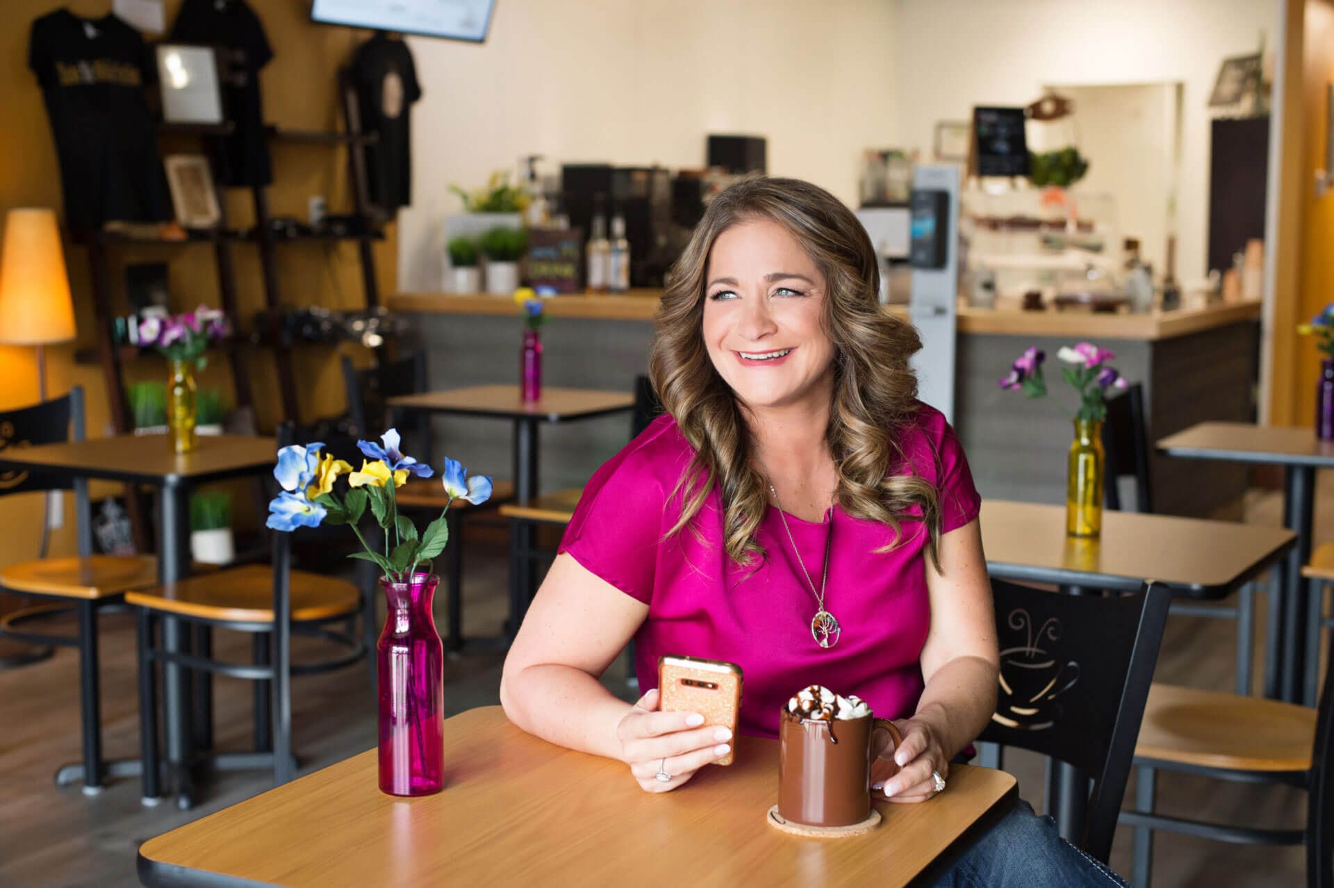 Woman smiling at a café table, holding a cup in a cozy coffee shop with flowers and mugs.