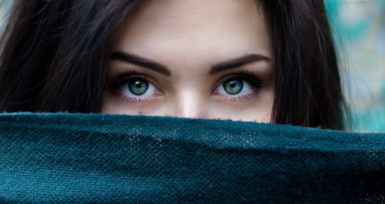 Close-up of a person peering over a teal scarf, with striking blue-green eyes and dark hair