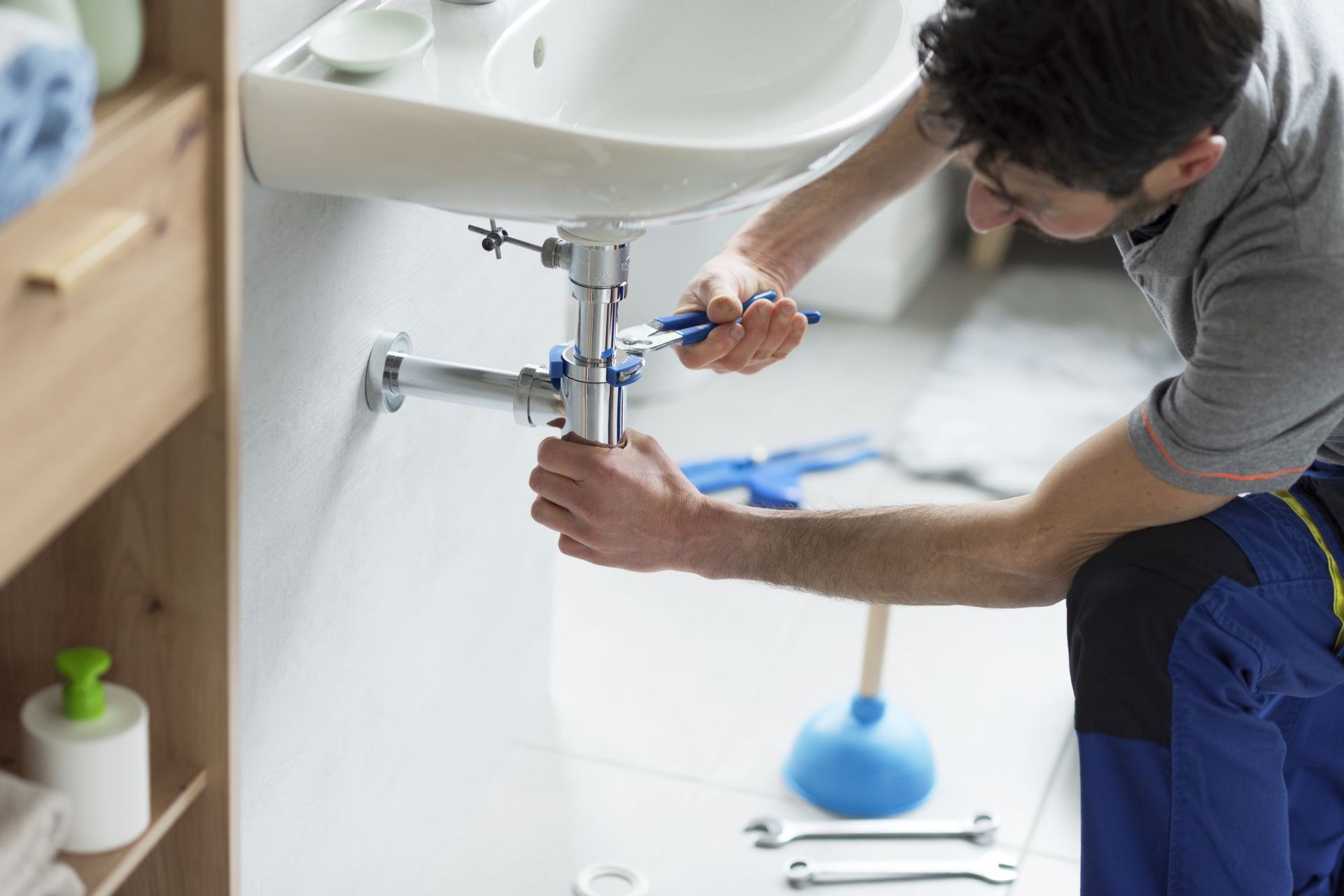 Man tightening pipes under a bathroom sink with a wrench, tools scattered nearby.