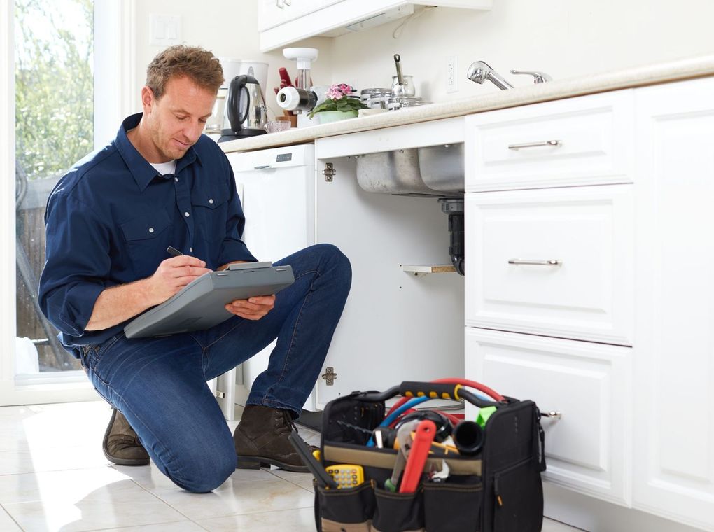 Plumber kneeling under a kitchen sink, inspecting plumbing with a tablet beside a toolbox.