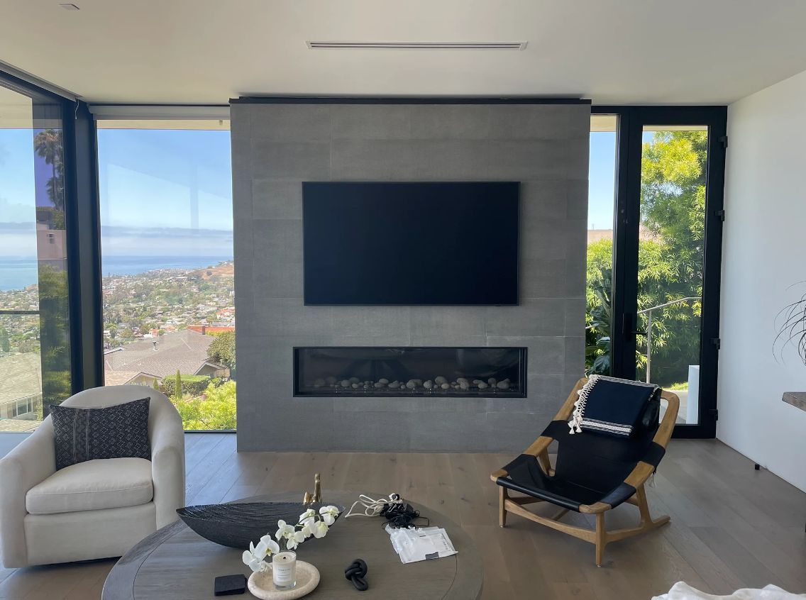 Modern living room with a wall-mounted TV, fireplace, large windows, and gray and black seating.