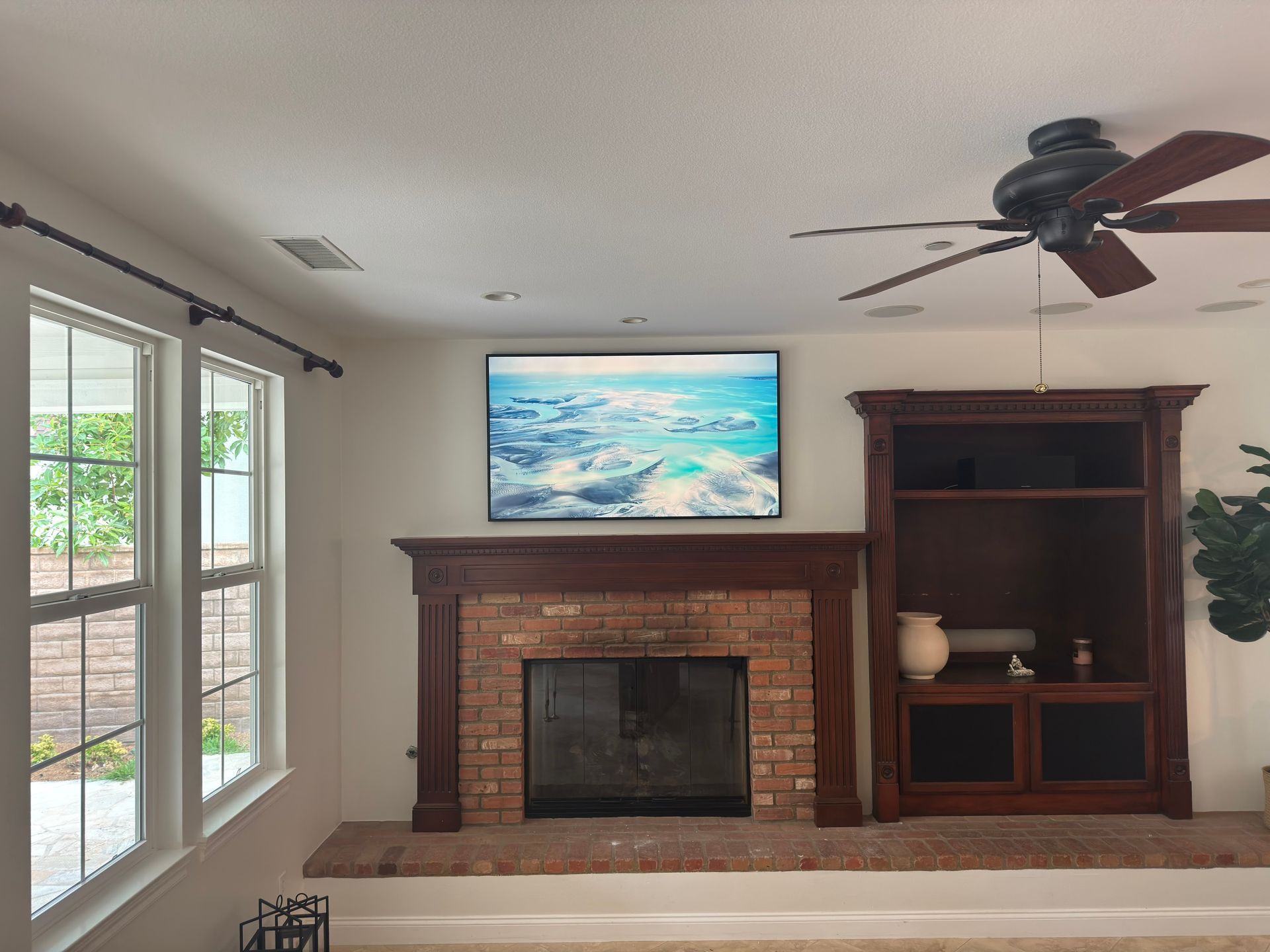 Living room with brick fireplace, wall-mounted TV, built-in shelves, ceiling fan, and large windows