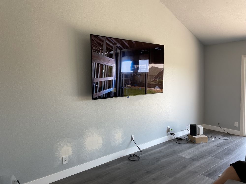 Wall-mounted TV in a sparse living room with light gray walls and wood flooring, with a few items on the floor.