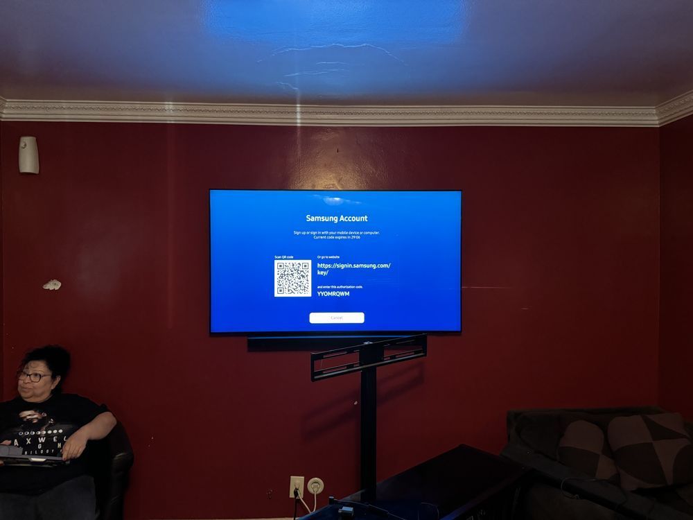 Living room with a wall-mounted TV on a stand showing a blue setup screen, and a seated person at left.