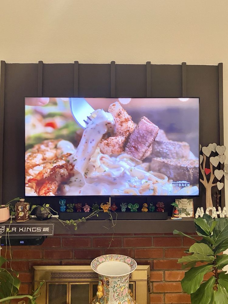 TV above a brick fireplace showing a close-up of grilled food, with plants and decor below