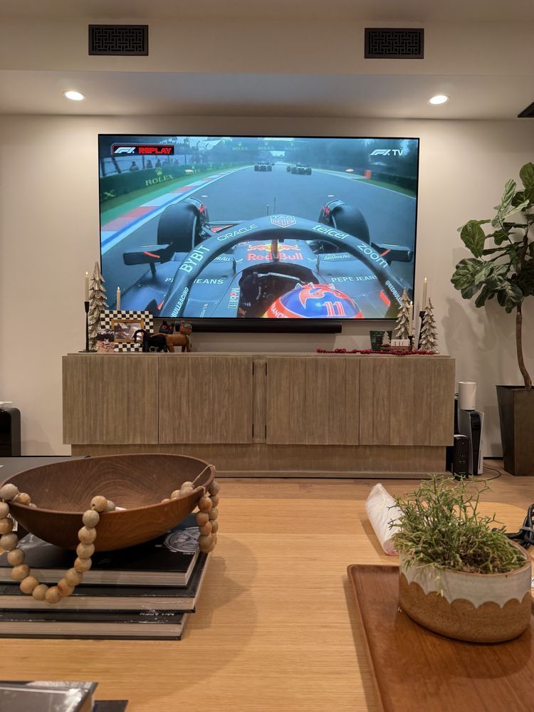Living room with large TV showing a Formula 1 race, wood console, plants, and a cozy chair in the foreground